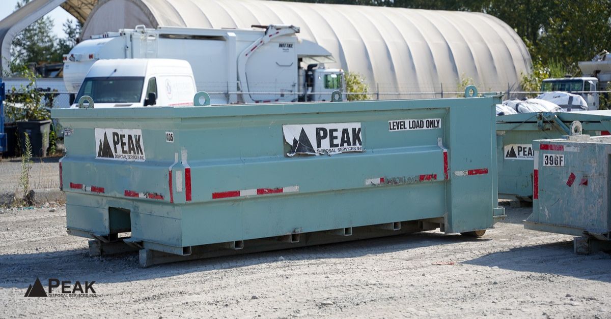 Large Peak Disposal dumpster in a gravel yard with company trucks and industrial buildings in the background