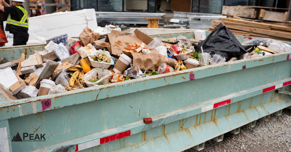 Construction dumpster filled with food scraps, takeout containers, coffee cups, and mixed building debris onsite.