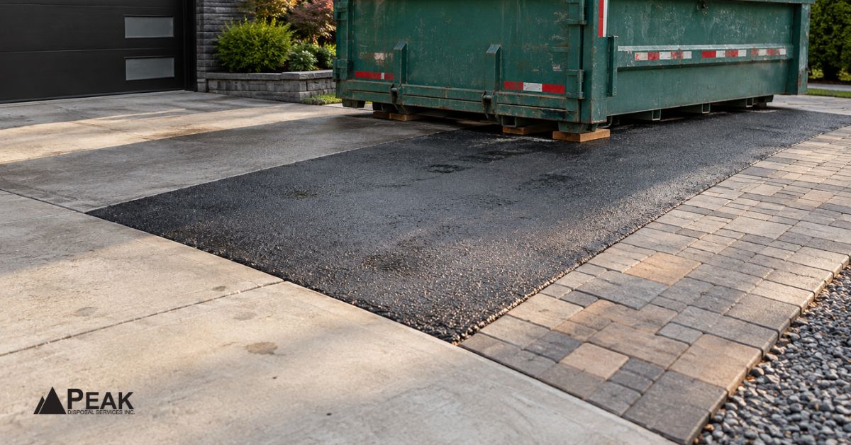 Roll-off bin sits across concrete, asphalt, pavers, and gravel driveway surfaces at a residential property.