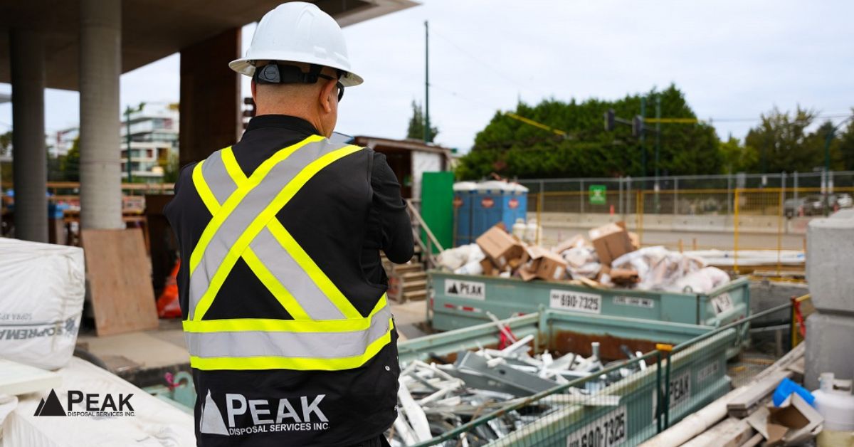 Peak Disposal worker oversees construction waste sorting with roll-off bins at a busy jobsite nearby.